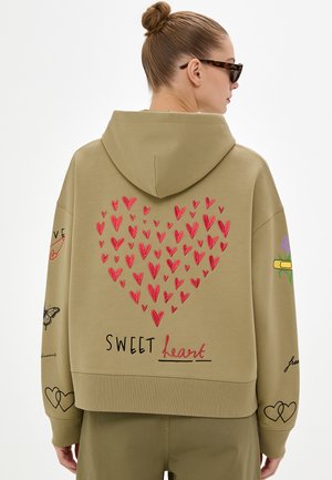 Donna che indossa una felpa con cappuccio color kaki con motivi a cuore rossi che formano un grande cuore e la parola "SWEET heart" sulla schiena, oltre a vari disegni sulle maniche.