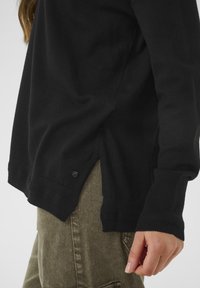 Maglione nero a maglia con polsini a coste, spacco laterale e dettaglio con bottoni. Il materiale appare morbido con una texture liscia, abbinato a pantaloni verde oliva.