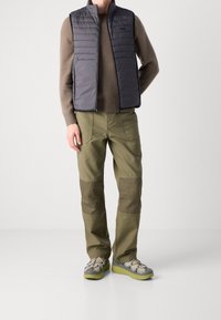 Grå vadderad väst över en brun stickad tröja, i kombination med gröna cargobyxor med mörkare lappar, och grå sneakers med gröna sulor.