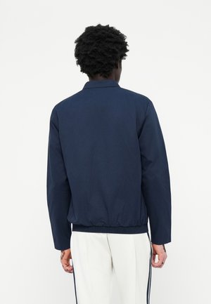Personne aux cheveux bouclés portant une veste bleu marine et un pantalon blanc avec des bandes latérales noires, debout de dos sur un fond uni.