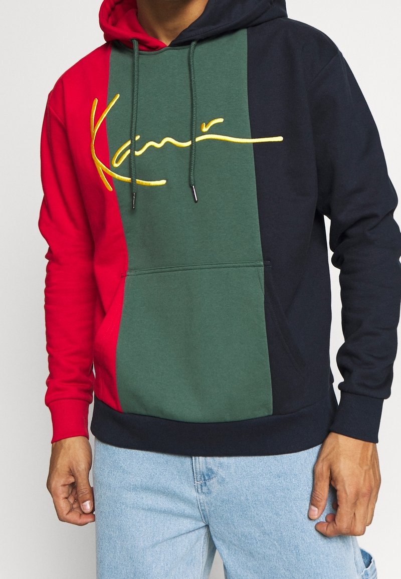 Homme portant un sweat à capuche tricolore rouge, vert et marine avec un logo jaune en cursive sur la poitrine et un jean bleu clair.