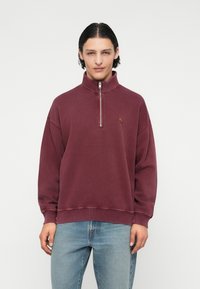 Sweatshirt bordeaux avec un col haut, une fermeture éclair quart de devant et un logo brodé sur la poitrine gauche. Tissu doux et décontracté.