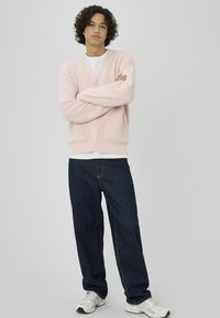 Cardigan rose pâle côtelé avec col en V et deux poches avant, associé à un pantalon en denim bleu foncé et des baskets blanches.