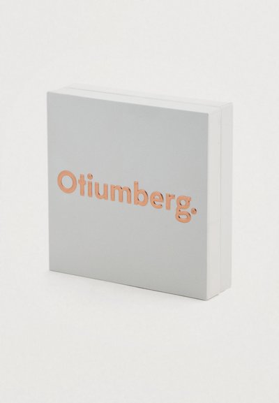 Τετράγωνο κουτί ανοιχτού γκρι χρώματος με υφή, που φέρει τη λέξη "Otiumberg." σε ροζ χρυσό, τοποθετημένη στο κέντρο της πρόσοψης.
