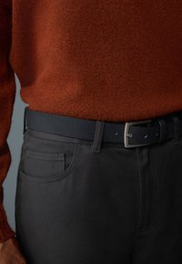 Czarny pas materiałowy o teksturowanym wykończeniu, z metalową klamrą. Noszony na ciemnoszarych spodniach i pomarańczowym, teksturowanym swetrze.