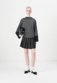 Blusa oversized listrada em preto e branco com mangas largas, combinada com uma saia plissada preta e mocassins pretos com laços. Pequena bolsa texturizada.