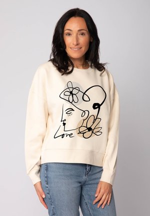 Sweatshirt couleur crème avec un dessin en ligne noire représentant un visage et des fleurs, avec des poignets côtelés et une coupe décontractée.