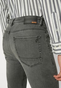 Gros plan sur la poche arrière d'un jean gris avec une étiquette en cuir noir affichant "BOSS" en orange, porté avec une chemise blanche à rayures grises.