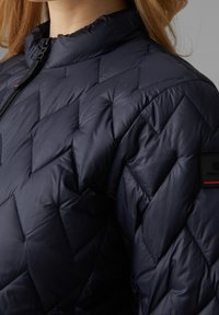 Tmavě modrá péřová bunda s diamantovým prošitím, zapínáním na zip, stojáčkem a logem na rameni. Jemná, lehká textura.