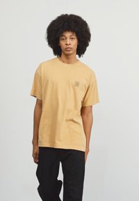 Camiseta de algodón color tostado con mangas cortas, cuello redondo y un pequeño parche con logo en el lado izquierdo del pecho. Ajuste holgado y diseño sencillo.