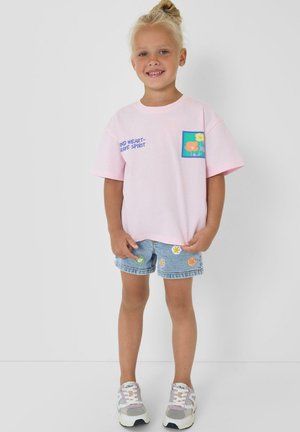 Jeune fille debout et souriante, portant un T-shirt rose à imprimé floral et un short en denim bleu décoré de fleurs brodées.