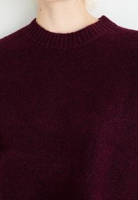 Pull col rond bordeaux avec col côtelé, fabriqué en tissu à texture douce, présentant une coupe décontractée et un design minimaliste.