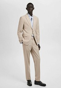 Beżowy garnitur o dopasowanym kroju, jednorzędowa marynarka, szpiczasty kołnierz, w zestawieniu z jasnoniebieską koszulą i czarnymi skórzanymi butami.