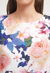 Bloemenprint op stof met een witte achtergrond, featuring grote roze pioenrozen, kleinere bloesems en groene bladeren in een levendige print.