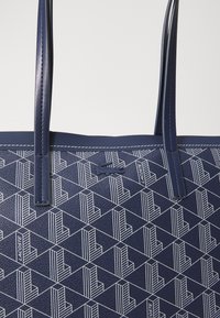 Mornarsko modra torba z teksturirano površino, ki ima geometrijski beli vzorec, dve dolgi ročaji in majhno logotipno oznako.