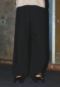 Pantalons larges noirs en tissu doux, offrant une coupe décontractée et de légères plis. Assortis avec des chaussures noires sur une surface texturée.