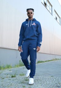 Kraljevo modra kapuca in hlače s belimi akcenti in Adidas logotipom, z vezeno črko, v kombinaciji z belimi športnimi copati in sončnimi očali.