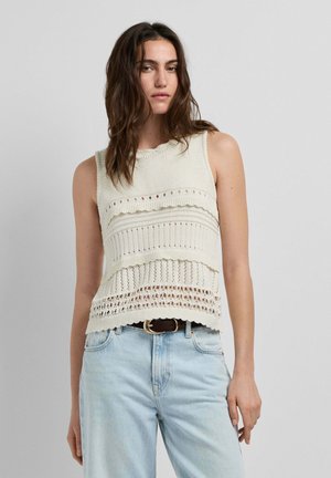 Mujer con cabello castaño ondulado, vistiendo un top de punto blanco sin mangas con patrones y jeans de cintura alta azul claro con un cinturón negro.