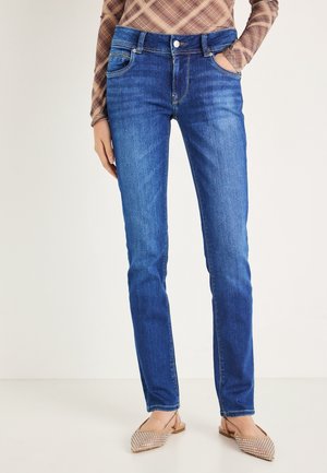 Frau trägt blaue Jeans, beige spitze Ballerinas und ein braunes kariertes Langarmshirt, steht mit einer Hand in der Tasche.