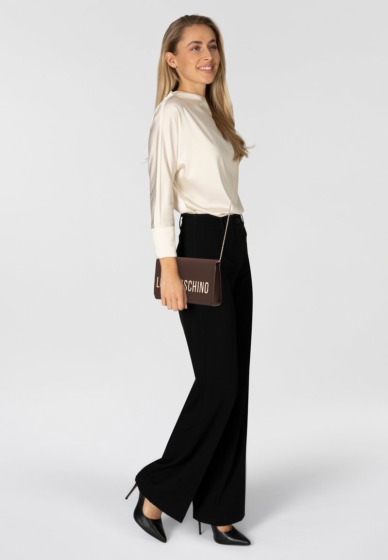Eine Frau trägt eine crèmesatinierte Bluse, schwarze hochtaillierte, weite Hosen, schwarze spitze Absatzschuhe und hält eine braune Moschino-Clutch.