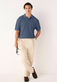 Cămașă polo texturată, de culoare albastră, cu guler, mâneci scurte și trei butoni, asortată cu pantaloni bej deschis și pantofi slip-on maro.