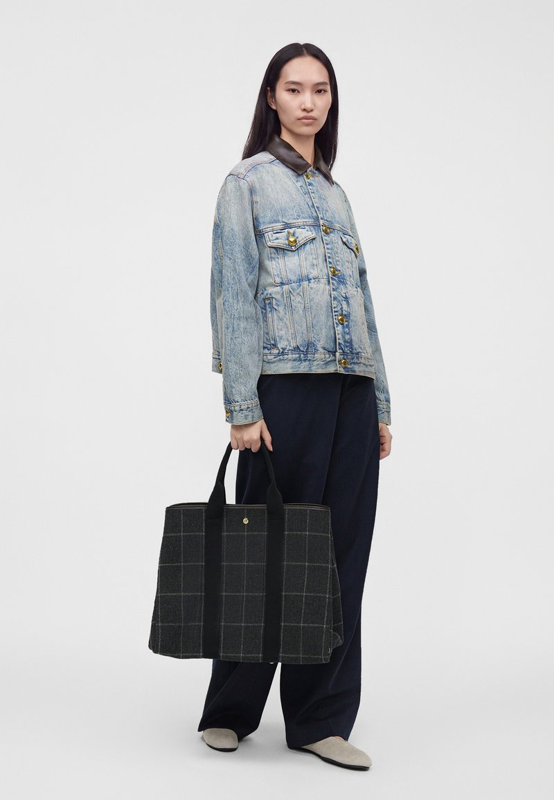 Femme portant une veste en jean claire avec un col foncé, un pantalon large noir, des chaussures beiges, tenant un grand sac cabas noir à carreaux par les poignées.