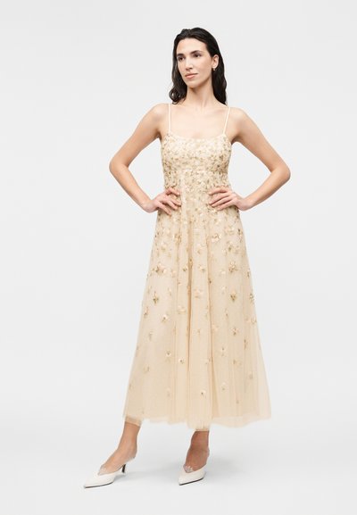 Γυναίκα στέκεται με τα χέρια στη μέση, φορώντας ένα μπεζ αμάνικο φόρεμα με λουλουδένια κέντημα και λευκά μυτερά τακούνια πάνω σε λευκό φόντο.