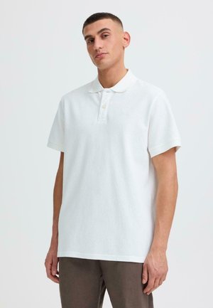 Jonge man met kort haar, gekleed in een wit poloshirt met korte mouwen en een bruine broek, staand tegen een eenvoudige lichte achtergrond.