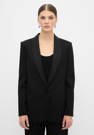Schwarzer Blazer mit Nadelstreifen, satiniertem Revers, einem Knopfverschluss und zwei vorderen Taschen, getragen über einem schwarzen gerippten Shirt.