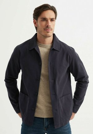 Veste en coton marine avec boutonnage, deux poches avant et un col. Portée sur un pull beige clair, associée à un jean bleu.