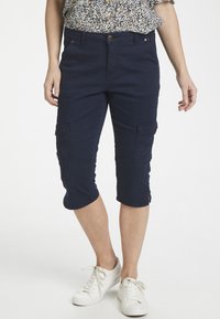 Marinblå capri cargobyxor med fickor, slim fit och dragkedjedetaljer vid fållen, tillsammans med vita sneakers. Lätt tygkänsla.