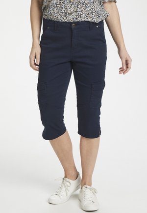 Pantalons cargo capri bleu marine avec poches à rabat, coupe slim et accents zippés au bas, associés à des baskets blanches. Texture de tissu légère.