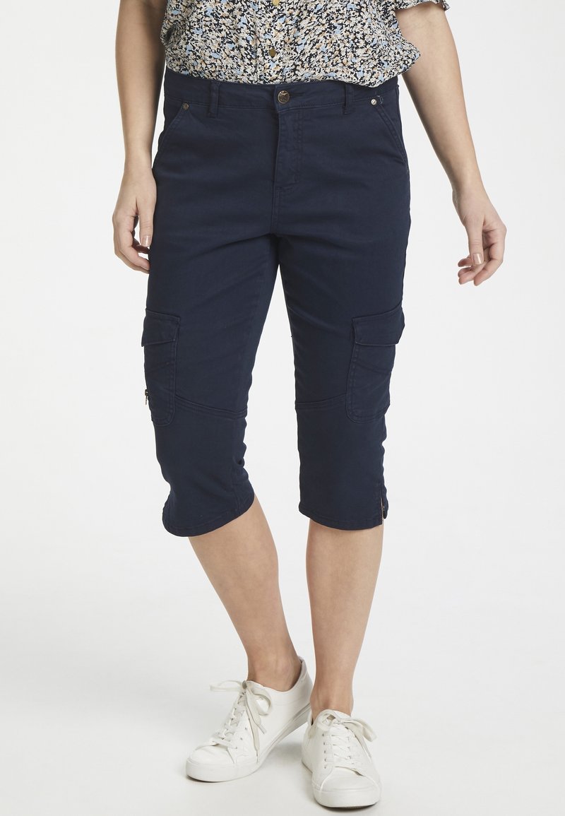 Marinblå capri cargobyxor med fickor, slim fit och dragkedjedetaljer vid fållen, tillsammans med vita sneakers. Lätt tygkänsla.