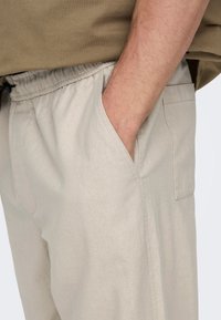 Beżowe bawełniane spodnie z elastycznym paskiem, bocznymi kieszeniami i fakturowanym wykończeniem. Praktyczny fason odpowiedni na codzienne noszenie.