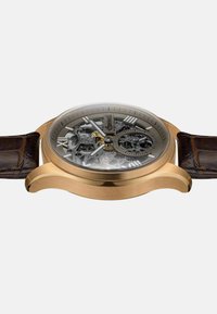 Montre automatique avec un boîtier en bronze, cadran transparent affichant des mécanismes complexes, et un bracelet en cuir marron avec texture crocodile.