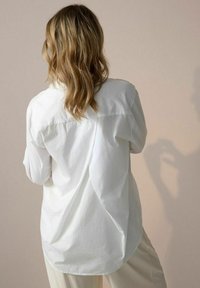 Camicia bianca a pulsante con una vestibilità comoda, presenta un orlo arrotondato e pieghe posteriori; indossata sopra pantaloni beige, il tessuto strutturato è visibile.