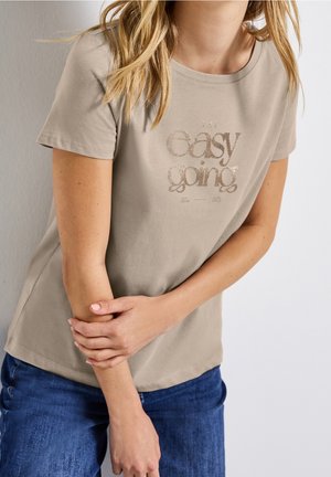 Kvinde iført beige t-shirt med teksten "The easy going", holder hendes venstre underarm, iført blå jeans mod en ensfarvet baggrund.