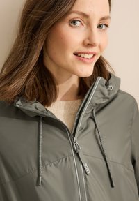 Giacca leggera grigia con cappuccio, dotata di una zip frontale e cordino. Texture liscia e design minimalista, adatta per un abbigliamento casual.
