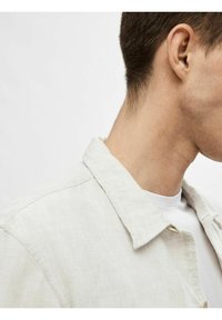 Lichtbeige linnen shirt met een kraag, die textuur en zachte stof toont. Daaronder is een wit T-shirt gedeeltelijk zichtbaar.