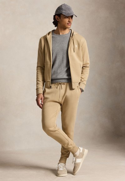 Homme portant un sweat à capuche beige zippé, un jogging assorti, un pull gris, une casquette grise et des baskets beiges, debout, jambes croisées sur fond neutre.