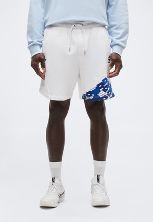 Mees, kes kannab valgeid spordipükse sinise geomeetrilise mustriga vasakul jalal, valgeid sokke, valgeid tossusid ja helesinist dressipluusi.