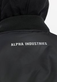 Zwarte jas met geribde kraag en witte tekst "ALPHA INDUSTRIES" gedrukt over de bovenrug, gedragen over een zwarte hoodie.