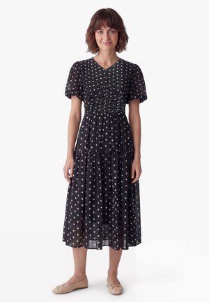 Woman with short brown hair wearing a black midi dress with white floral print and puff sleeves, standing with beige ballet flats on white background.