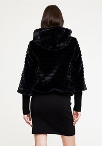 Svart faux fur kort jacka med huva, med vertikala ränder och långa svarta ribbade ärmar. Buren över en figurnära svart klänning.