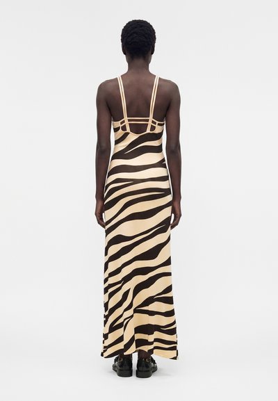 Γυναίκα γυρισμένη πλάτη φοράει μακρύ μπεζ φόρεμα με μαύρες ζεβρέ ρίγες και λεπτούς διπλούς ιμάντες στους ώμους, συνδυασμένο με μαύρα παπούτσια.