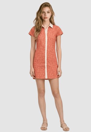 Mujer joven de pie, vistiendo un vestido floral naranja de manga corta con cuello blanco y franja frontal, combinado con sandalias beige.