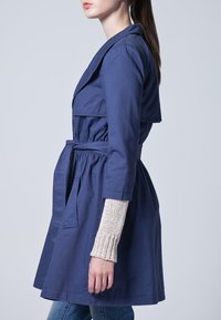 Granatowy płaszcz typu trencz z paskiem w talii, z podwójnym kołnierzem i rękawami o długości trzech czwartych. Widoczna teksturowana, kremowa warstwa podkoszulka.