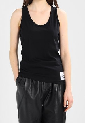 Schwarzes ärmelloses Tanktop aus Baumwolle mit rundem Ausschnitt und einem kleinen weißen Etikett unten links. Kombiniert mit schwarzen Lederhosen.