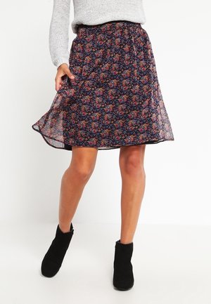 Woman wearing a navy floral skirt, gray knit sweater, and black ankle boots, standing on a white background.