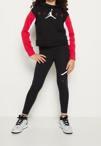 Bambino che indossa un hoodie nero con maniche rosse e bianche, leggings neri con logo e sneakers alte bianche con lacci neri, in piedi con le mani sui fianchi.